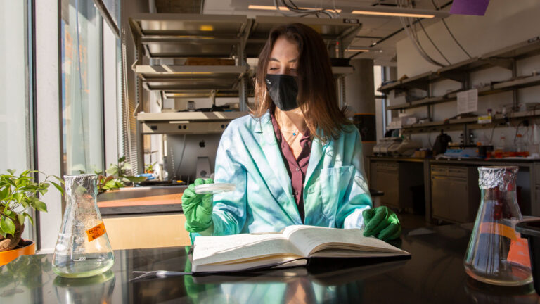 A FURI student reads a book and looks at a petri dish in a lab.