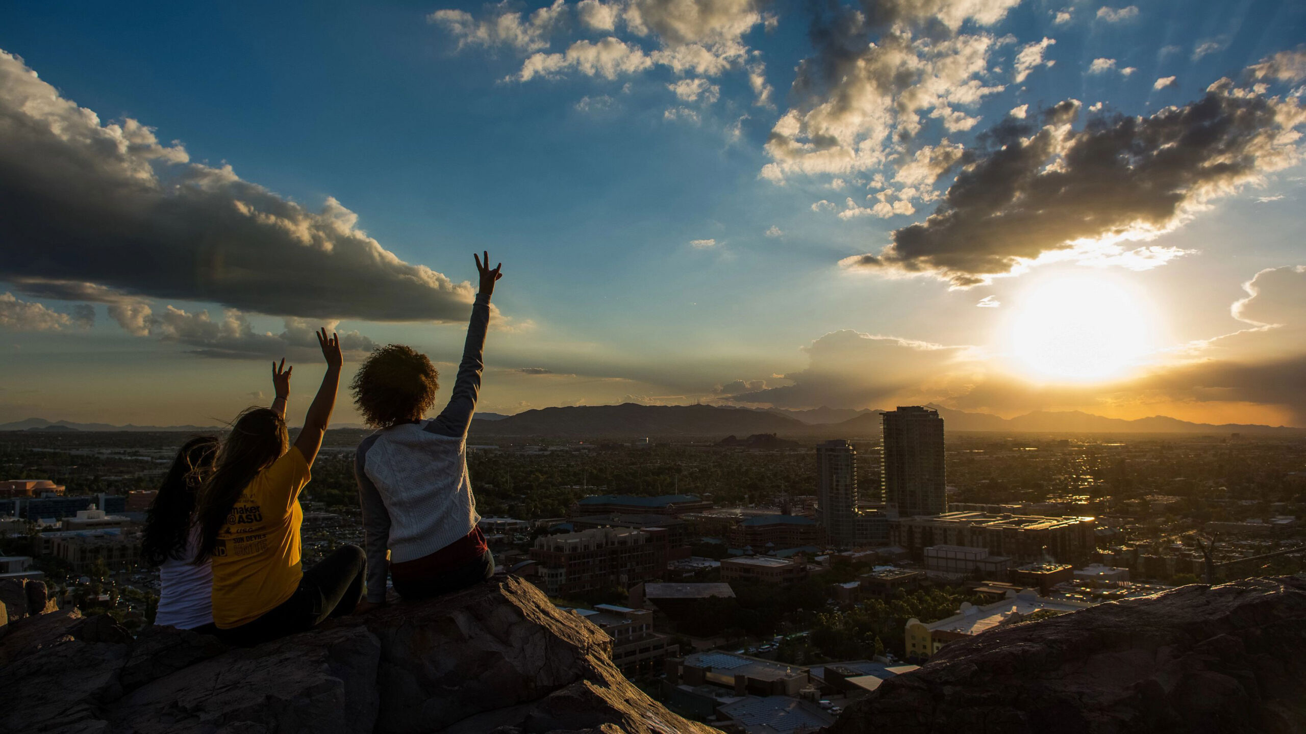 Two student sitting on the summit of A Mountain making the fork hand signal while looking down at the ASU Tempe campus.