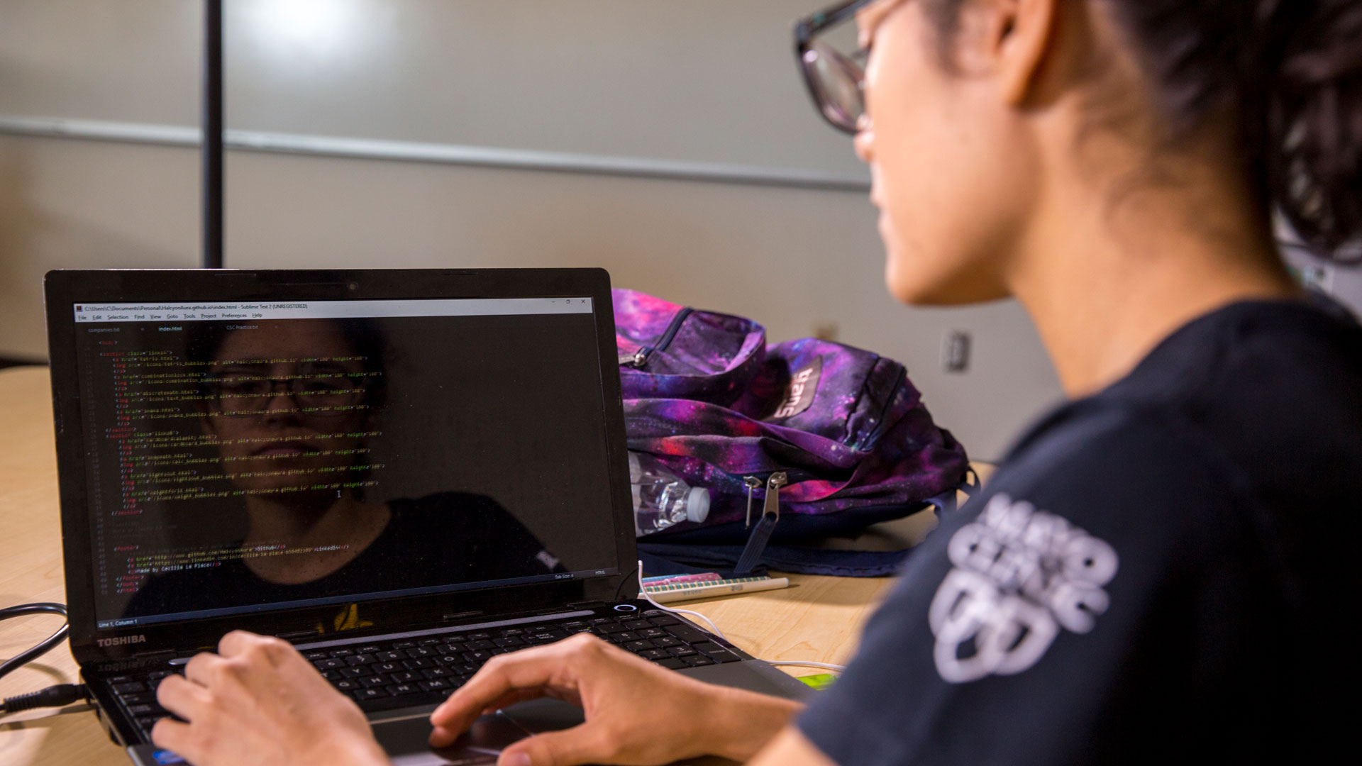 A photo of a student writing code with their reflection in the laptop screen.