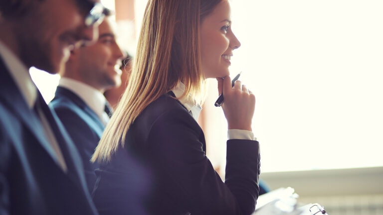 A woman in a business suit with a clipboard sits leaning forward.