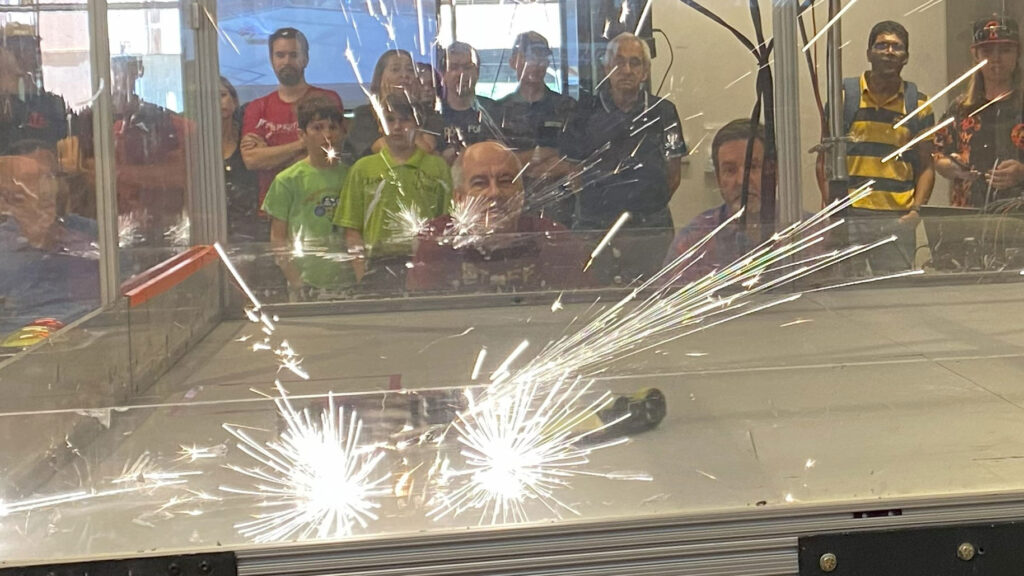 Two robots clash in a shower of sparks at a robot combat event, with spectators cheering them on in the background.