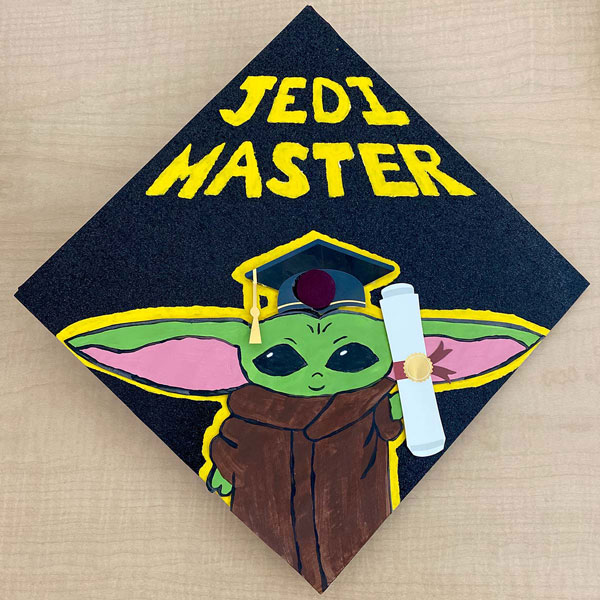 A decorated mortarboard featuring a baby yoda with a graduation mortarboard cap and a diploma and the words "Jedi Master."
