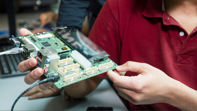 A circuitboard is held by a guy in a red shirt.