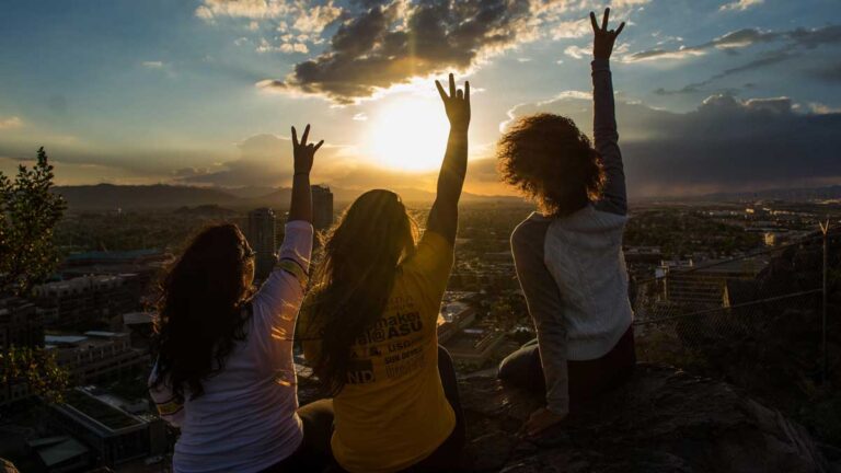 Three students sit on A Mountain and give the fork hand signal with a sunset in the background.