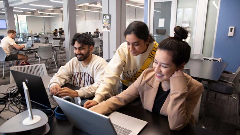 Three young people work together on laptops as one of them points at something on-screen over another's shoulder.